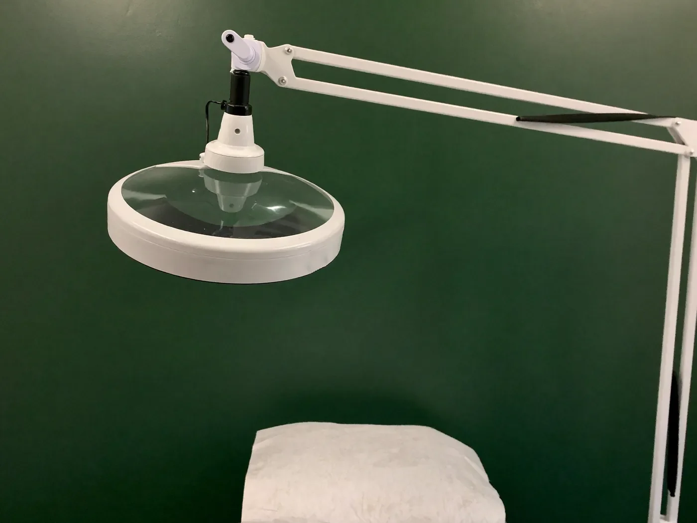 Sala de atendimento: a lupa de bancada branca, de braço articulado e apagada, sobre a cabeceira da maca coberta com papel, contra a parede verde