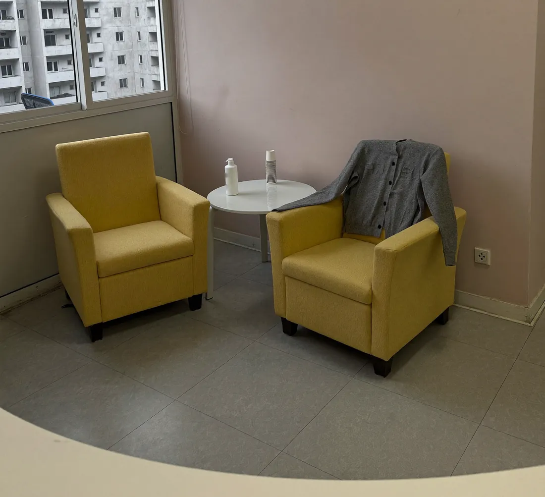 Canto de espera com duas poltronas amarelas, um casaco cinza largado numa delas, mesa branca com copos de papel e janela para um prédio residencial