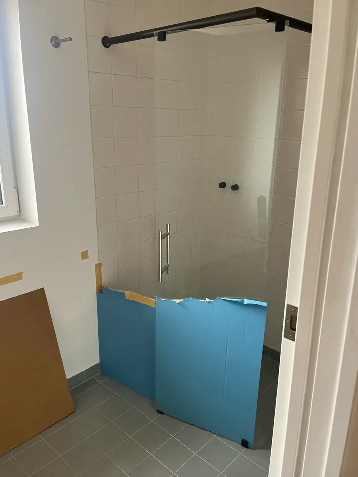 Box de canto de vidro incolor recém-instalado num banheiro de azulejo branco: trilho tubular preto, roldanas aparentes, puxador de inox e chapas azuis de proteção na parte de baixo das folhas.