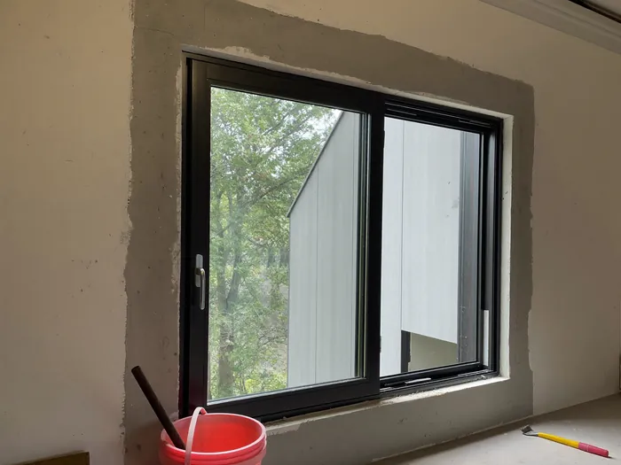 Janela de correr de alumínio preto recém-instalada, com a massa cinza do arremate ainda sem pintura em volta e um balde vermelho embaixo.