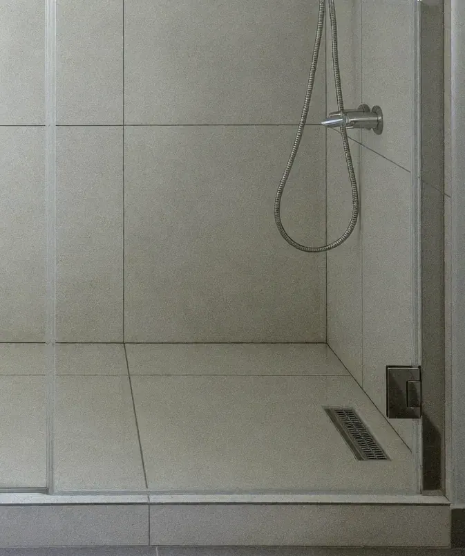 Canto de um box de vidro incolor num banheiro de porcelanato cinza: a ducha com mangueira, o ralo linear e a dobradiça presa na parede.