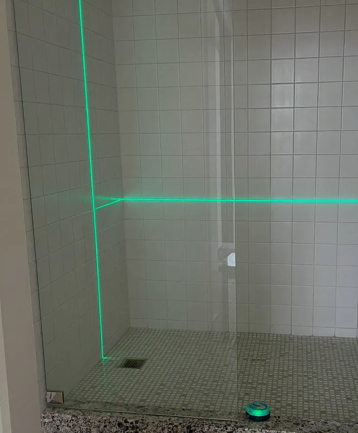 Vão de box num banheiro de azulejo branco, com a folha fixa já no lugar à esquerda. Um nível a laser no chão projeta uma linha verde em pé na parede da esquerda e outra deitada atravessando a parede do fundo.