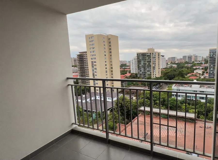Vista de uma sacada no 14º andar: a parede branca à esquerda, o guarda-corpo de alumínio e, lá fora, prédios residenciais, telhados vermelhos e árvores sob o céu nublado.
