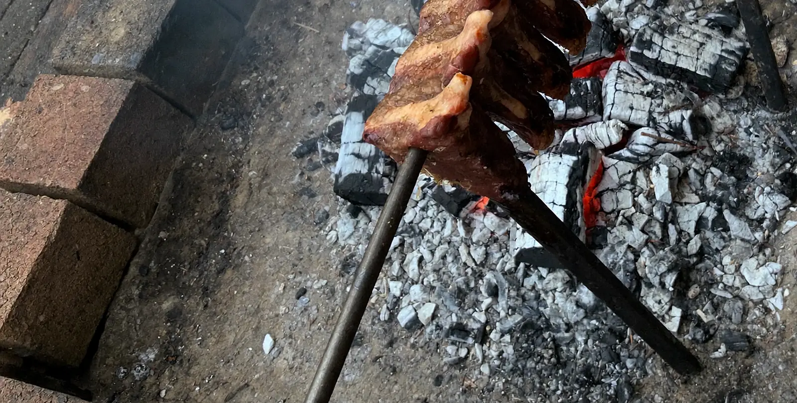 A ponta de uma manta de costela no espeto de ferro, sobre a brasa do fogo de chão: os carvões cobertos de cinza branca, com o vermelho aparecendo nas rachaduras.
